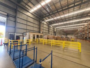 Interior of a large warehouse with yellow safety barriers, stacked cardboard boxes, and metal shelves.