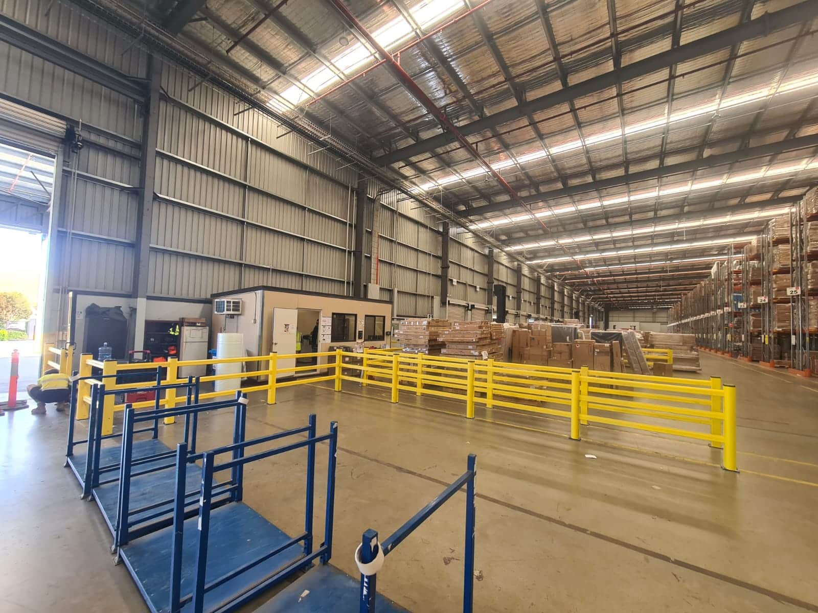 Interior of a large warehouse with yellow safety barriers, stacked cardboard boxes, and metal shelves.