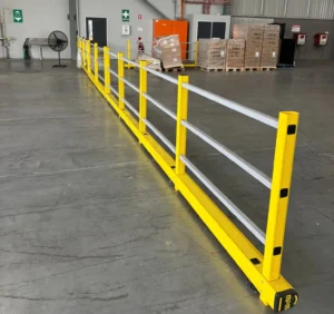 Yellow safety barrier with silver horizontal rails installed in a warehouse to separate pedestrian walkway from storage area with pallets.